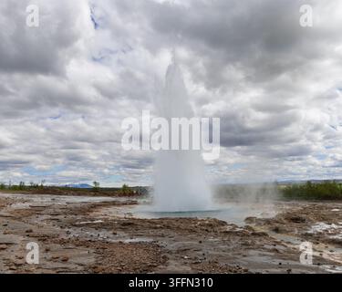 Der Strokkur-Geysir bricht aus einem blauen Becken in Haukadalur, Island, hervor, mit einer dichten weißen Wassersäule, die unter dramatischen geschichteten Sommerwolken zentriert ist. Stockfoto