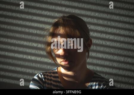 Trübsinnige junge Frau in gestreiftem Hemd nach vorne gerichtet mit dramatischem Sonnenlicht und blinden Schatten, die sich über das Gesicht und den texturierten Wandhintergrund ziehen. Stockfoto