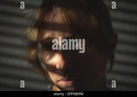 Nahaufnahme-Porträt einer nachdenklichen jungen Frau mit dramatischem Sonnenlicht und Blenden-Schatten über dem Gesicht, die ihr ausdrucksstarkes Auge hervorhebt. Stockfoto