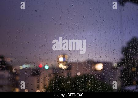 Blick durch eine regennasse Fensterscheibe in der Dämmerung oder im Zwielicht, mit schweren Wassertropfen auf dem Glas und einer verschwommenen Stadtsilhouette Stockfoto