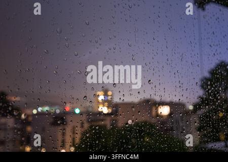 Nahaufnahme eines Fensters, das während eines Abendregens mit Regentropfen bedeckt ist und einen weichen städtischen Hintergrund mit fernen Gebäudeleuchten und dunkler Silhouette einrahmt Stockfoto