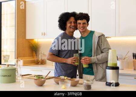 Afrikanische männliche Partner stehen, lächeln und halten grüne Smoothies neben dem Mixer in der Küchenstube zu Hause Stockfoto