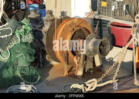 Verwittertes Fischerbootdeck mit rostiger Winde, grünen Netzen, aufgerollten Seilen und maritimer Ausrüstung, Nahaufnahme-Detail, das abgenutztes Metall, Ketten, Rollen und Arbeitsgeräte zeigt Stockfoto