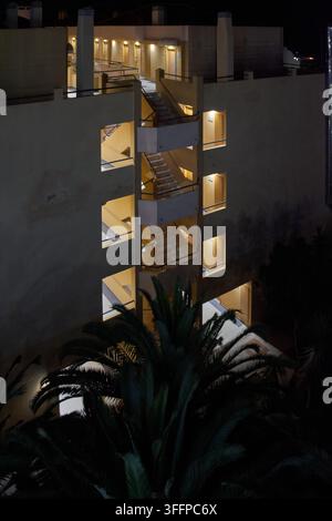 Außenperspektive eines mehrstöckigen Wohnkomplexes bei Nacht mit warmer Innenbeleuchtung und Palmen Stockfoto
