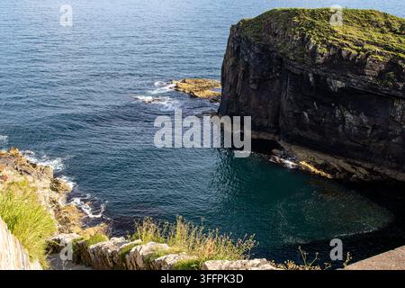 Schroffe Klippenwand trifft auf tiefblaues Meer mit grüner, grasbewachsener Spitze und felsiger Küstenlinie an den Whaligoe Steps in Schottland, UK. Stockfoto
