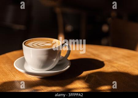 Nahaufnahme einer weißen Kaffeetasse mit Blatt-Latte-Art auf einem hölzernen Cafétisch, beleuchtet von warmem Sonnenlicht und dramatischen Schatten in einem gemütlichen Kaffeehaus Stockfoto