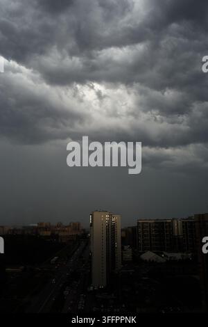 Dramatische dunkle Sturmwolken sammeln sich über einer dichten städtischen Skyline, mit einer hellen Lücke im bedeckten Himmel und heftigem Regen, der auf Hochhauswohnungen herabnimmt Stockfoto