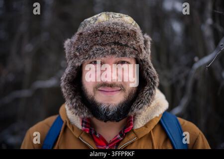 Nahaufnahme eines lächelnden bärtigen Mannes, der im Winter im Freien in die Kamera schaut, mit einem Pelz-Ushanka-Hut, brauner Jacke, kariertem Hemd und Rucksack Stockfoto