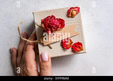 Nahaufnahme einer Hand, die eine handgemachte Geschenkschachtel hält, die mit einer getrockneten roten Rose, getrockneten Rosenblättern, Schnur und einem Kraftpapiertag verziert ist, der mit dem Wort gestempelt ist Stockfoto