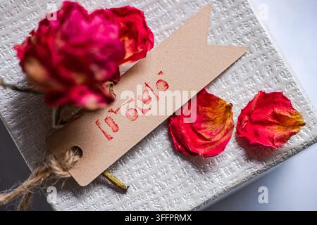 Nahaufnahme von getrockneten roten Rosenblättern und einer Rose, die auf einer strukturierten weißen Geschenkschachtel ruht, mit einem Kraftpapieranhänger, der mit dem Wort „Liebe“ gestempelt ist und mit Nat gebunden Stockfoto