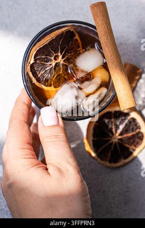 Überkopf-Nahaufnahme einer Hand, die gewürzten Eiskaffee in einem Glas mit Eiswürfeln, Zimtstange und getrockneter Orangenscheibe auf einer grauen Steinfläche hält. Stockfoto