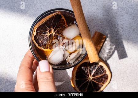Oberblick einer Hand, die eisgekühlten gewürzten Kaffee in einem Glas hält, garniert mit einem Zimtstößel und einer getrockneten Orangenscheibe auf einer sonnenbeschienenen grauen Oberfläche. Stockfoto