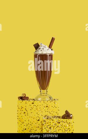 Reicher Schokoladenmilchshake mit Schlagsahne, Schokoladenstücken und einer Waffelrolle, präsentiert in einem hohen Glas auf einem hellgelben, mit Streuseln bedeckten Untergrund Stockfoto