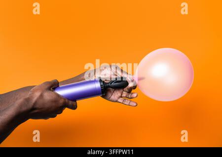Nahaufnahme von dunkelhäutigen Händen, die mit einer lila Handpumpe einen rosa Ballon gegen einen lebhaften orangefarbenen Hintergrund aufblasen und damit eine verspielte Feierlichkeit schaffen Stockfoto