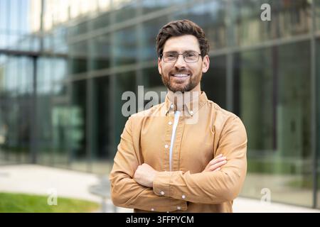 Junger Geschäftsmann mit Brille und braunem Hemd, die Arme verschränkt, der Selbstvertrauen und Professionalität vor einem modernen Glasbürogebäude ausdrückt Stockfoto