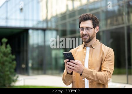 Junger Mann mit Brille lächelt und schaut auf sein Mobiltelefon, verbindet sich mit digitalen Inhalten und sozialen Medien im Freien in einer modernen Stadtumgebung Stockfoto