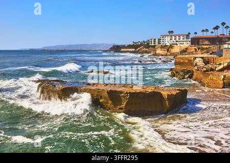 Helles Sonnenlicht badet die raue Küstenlinie der Sunset Cliffs in San Diego, Kalifornien, während Meereswellen gegen große Felsen und Strandgebäude schlagen Stockfoto