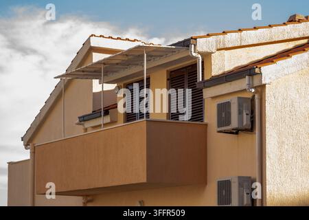 Eine beige Wohnhausfassade zeigt einen Balkon, geschlossene Fensterläden und montierte Klimaanlagen. Stockfoto