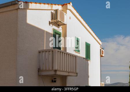 Ein weißes Stuckhaus mit grünen Fensterläden, einem kleinen Balkon und wandmontierten Klimaanlagen unter einem blauen Himmel. Stockfoto