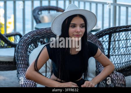 Junge Frau mit einem weißen Hut sitzt auf einem Korbstuhl in der Nähe des Wassers. Stockfoto