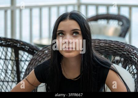 Eine junge Frau mit langen dunklen Haaren sitzt in einem Korbstuhl und schaut nachdenklich nach oben. Stockfoto
