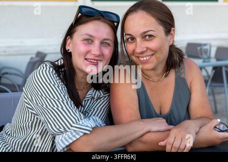 Zwei Frauen lächeln, während sie an einem Tisch in einer Außengastronomie-Einstellung zusammenlehnen. Stockfoto