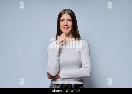 Porträt einer nachdenklichen jungen Frau, die die Hand am Kinn hält, vor grauem Hintergrund Stockfoto