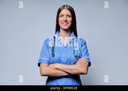 Selbstbewusste Krankenschwester in blauer Uniform mit Stethoskop, die mit verschränkten Armen vor grauem Hintergrund steht Stockfoto