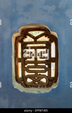 Verziertes hölzernes Fenstergeflecht am Hon-Chen-Schrein in Hue, Vietnam. Eine Detailaufnahme traditioneller architektonischer Schnitzereien, die in eine blau gestrichene Wand eingelassen sind Stockfoto