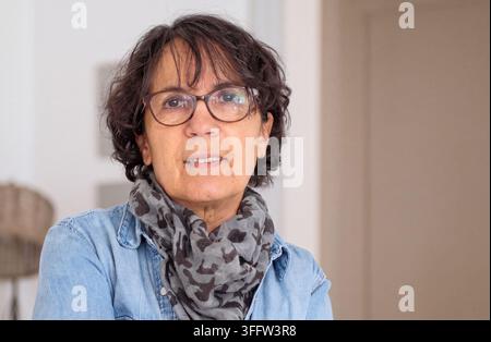 Nahaufnahme eines Indoor-Porträts einer mittelalten Frau mit kurzen dunklen lockigen Haaren und einer Brille, die ein Denimhemd und einen gemusterten Schal trägt und ruhig blickt Stockfoto