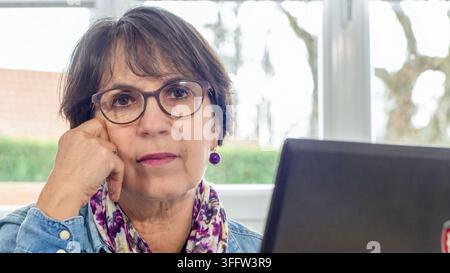 Nahaufnahme-Porträt einer ernsten Frau mittleren Alters mit Brille und einem bunten Schal, die sich neben einem Laptop auf ihre Hand stützt und remote arbeitet Stockfoto
