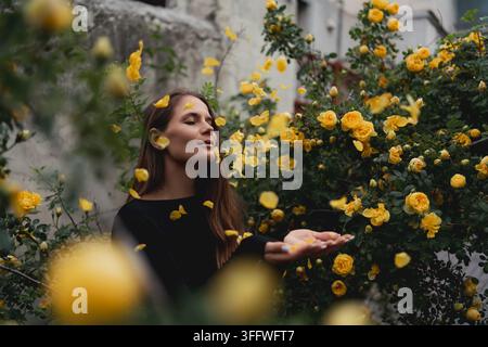 Junge Frau genießt fallende gelbe Rosenblätter in einer üppigen Sommergartenumgebung Stockfoto