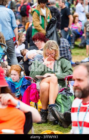 FRAU, DIE IN EINER FESTIVALTELLE EIN BUCH LIEST, REPORTAGE, GREEN MAN FESTIVAL 2015, BRECON, WALES, UK, 2015: Eine Frau mit Brille sitzt auf einem Klappstuhl und liest ein Buch inmitten einer geschäftigen Festivalmenge, während Kinder in der Nähe auf dem Gras sitzen. Foto: Rob Watkins. INFO: Das Green Man Festival findet jährlich im August in den Brecon Beacons statt. Es feiert alternative Musik, Kunst, Literatur, Wissenschaft und familienfreundliche Kultur in einer wunderschönen Berglandschaft. Das Event 2015 begann sonnig, wurde aber durch nächtlichen Regen an den letzten beiden Tagen schlammig. Stockfoto