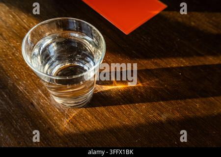 Klares Glas Wasser auf einem dunklen Holztisch, beleuchtet von warmem Sonnenlicht mit langen Schatten und einer roten Karte im Hintergrund. Stockfoto