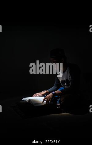 Asiatischer muslimischer Mann mit Peci-Hut, der den Heiligen Koran oder ein Buch liest, sitzt auf dem Boden bei gedämpfter Beleuchtung und dunklem schwarzem Hintergrund mit Kopierraum. Stockfoto