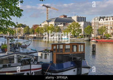 Amsterdam, Niederlande – 28. August 2026: Nahaufnahme traditioneller Wohnhausboote an der Amstel mit dem Royal Theatre Carre im Hintergrund am Stockfoto