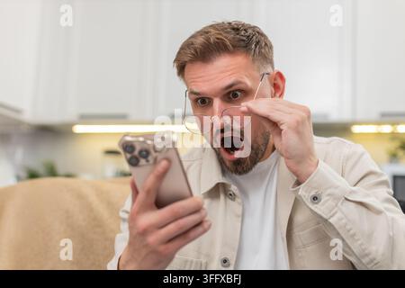 Ein junger Mann zu Hause liest mit einer Brille winzige Texte auf dem Smartphone und staunt über die Geschichte und Neuigkeiten. Der kaukasische Kerl auf dem Sofa sieht starke Aufmerksamkeit im Boten, zeigt Freude Erleichterung und helle Wow-Überraschung auf einmal Stockfoto