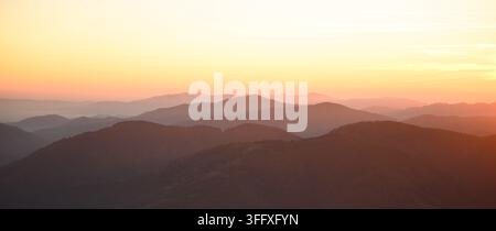 Schichtige Bergkämme glühen im warmen Licht bei Sonnenaufgang mit einem dunstigen goldenen Himmel. Minimale Panoramalandschaft mit atmosphärischen fernen Gipfeln Stockfoto