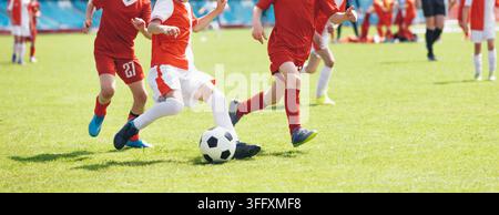 Junior-Fußballspieler in rot-weißer Uniform dribbelt den Fußball während eines Wettkampfs – Jugendfußball-Aktionsbild auf dem Rasenplatz Stockfoto