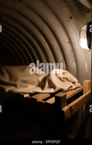 Inneneinrichtung eines rekonstruierten Luftschutzbunkers aus dem Zweiten Weltkrieg, der ein holzgerahmtes Bett mit einer gefalteten Wolldecke in einem gewölbten Wellblechbunker zeigt Stockfoto