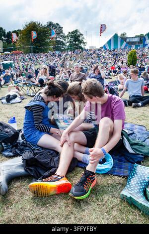 JUNGE FREUNDE TEILEN EIN BUCH AUF EINEM FESTIVAL, REPORTAGES, GREEN MAN FESTIVAL 2015, BRECON, WALES, UK, 2015: Eine Gruppe von vier jungen Freunden drängen sich auf einer Picknickdecke zusammen und blicken konzentriert auf ein Buch oder Programmheft, umgeben von einer großen Menschenmenge auf einem grasbewachsenen Hügel. Foto: Rob Watkins. INFO: Das Green Man Festival findet jährlich im August in den Brecon Beacons statt. Es feiert alternative Musik, Kunst, Literatur, Wissenschaft und familienfreundliche Kultur in einer wunderschönen Berglandschaft. Das Event 2015 begann sonnig, wurde aber durch nächtlichen Regen an den letzten beiden Tagen schlammig. Stockfoto