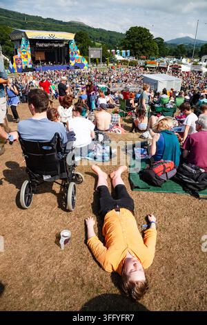 FESTIVALBESUCHER SCHLAFT AUF DEM GRAS, REPORTAGE, GREEN MAN FESTIVAL 2015, BRECON, WALES, UK, 2015: Eine Person in einem hellen orangefarbenen Hemd liegt rücklings tief schlafend auf dem trockenen Gras bei einem sonnigen Festival, mit einer Person im Rollstuhl und der großen Menge in der Nähe. Foto: Rob Watkins. INFO: Das Green Man Festival findet jährlich im August in den Brecon Beacons statt. Es feiert alternative Musik, Kunst, Literatur, Wissenschaft und familienfreundliche Kultur in einer wunderschönen Berglandschaft. Die Veranstaltung 2015 begann sonnig, wurde aber durch nächtlichen Regen an den letzten beiden Tagen schlammig. Stockfoto
