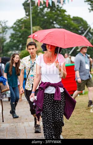 FRAU MIT ROTEM REGENSCHIRM UND GESICHTSBEMALUNG IM REGEN, REPORTAGE, GREEN MAN FESTIVAL 2015, BRECON, WALES, UK, 2015: Eine Frau mit Brille und rotem Puder im Gesicht hält einen roten Regenschirm, um sich vor dem Regen zu schützen, während sie auf einem Open-Air-Festival geht. Foto: Rob Watkins. INFO: Das Green Man Festival findet jährlich im August in den Brecon Beacons statt. Es feiert alternative Musik, Kunst, Literatur, Wissenschaft und familienfreundliche Kultur in einer wunderschönen Berglandschaft. Das Event 2015 begann sonnig, wurde aber schlammig durch nächtlichen Regen an den letzten beiden Tagen. Stockfoto