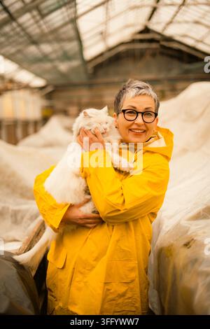 Zufriedene ältere Gärtnerin hält flauschige weiße Katze im Gewächshaus Stockfoto
