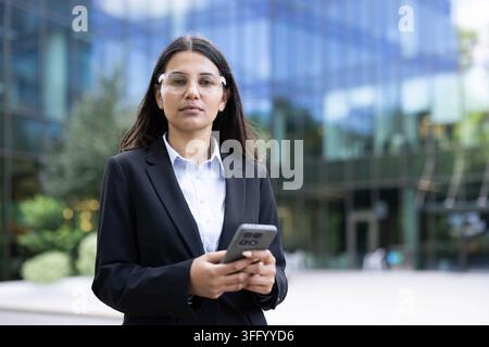 Geschäftsfrau mit Brille und maßgeschneidertem Anzug steht im Freien vor einem modernen Glasbürogebäude, hält ein Smartphone und schaut in die Kamera, vermittelt Selbstvertrauen und beruflichen Erfolg Stockfoto