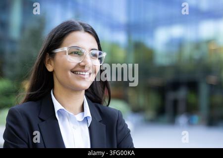Professionelle Geschäftsfrau mit Brille und Anzug, die selbstbewusst lächelt und nach vorne schaut, steht im Freien mit einem modernen Bürogebäude, das den Himmel im unscharfen Hintergrund reflektiert Stockfoto