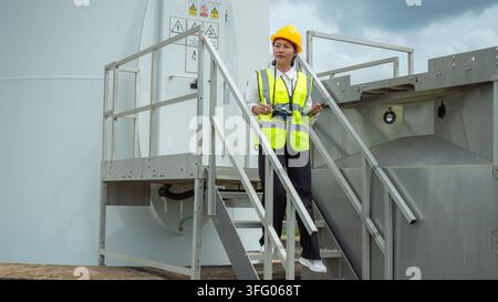 Asiatische Ingenieurin trägt einen gelben Sicherheitshelm und eine reflektierende Weste und hält ein digitales Tablet während der Inspektion einer Windturbine für erneuerbare grüne Energie Stockfoto