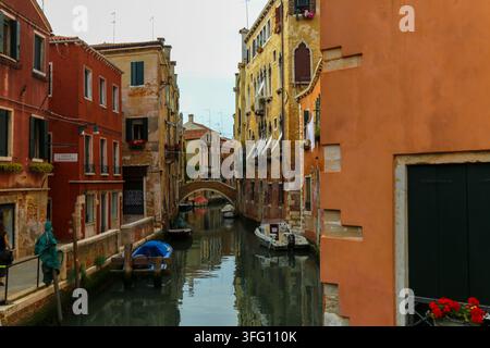 Ein ruhiger venezianischer Kanal mit farbenfrohen historischen Wohnhäusern, festgemachten kleinen Motorbooten und einer entfernten Steinbogen-Fußgängerbrücke in Venedig, Italien. Stockfoto