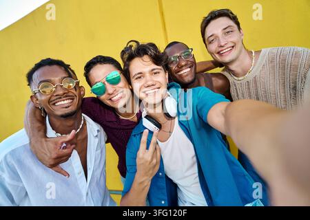 Gruppe glücklicher junger multikultureller Freunde, die in die Kamera lächeln, sich umarmen und einen Sommertag vor einer gelben Wand genießen Stockfoto
