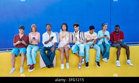 Gruppe multiethnischer junger Freunde, die sitzen, Smartphones halten und sich online verbinden vor einer lebendigen blau-gelben Wand Stockfoto